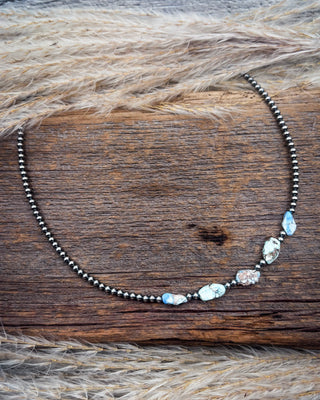 A beaded necklace with dark metallic beads and irregular light blue and white stones, displayed on a rustic wooden surface with dried grass.