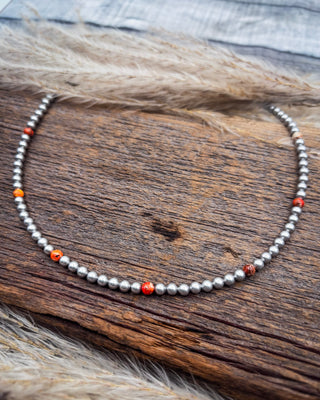 A beaded necklace made of small metallic silver beads with a few orange-red accent beads, displayed on a rustic wooden surface with dried grass.
