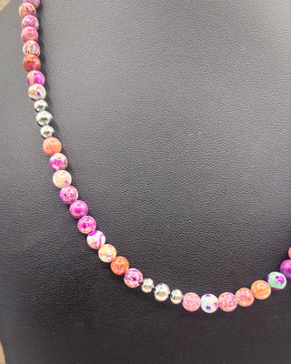 A beaded necklace with alternating pink marbled beads, orange marbled beads, and small metallic silver beads, displayed on a black textured surface.