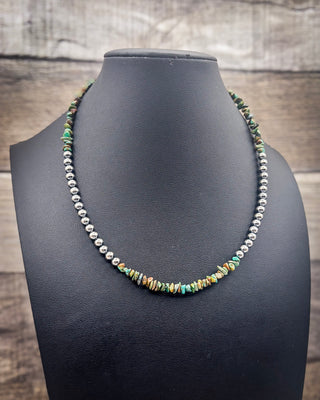 A beaded necklace displayed on a black jewelry bust, featuring a strand of silver beads and a central cluster of small multicolored beads with turquoise, green, orange, and gold tones, set against a rustic wood background.