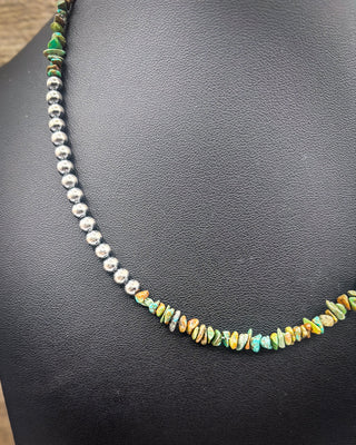 A beaded necklace displayed on a black textured jewelry bust. The necklace features a strand of polished silver beads on one side and a strand of small, irregular, multicolored stone chips on the other, with a small section of green stone beads at the top.