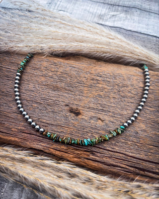 A beaded necklace with small metallic silver beads and a central cluster of irregular turquoise and brown stone beads, displayed on a rustic wooden surface with dried grass.