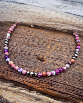 A beaded necklace made of round, polished gemstone beads in pink, purple, orange, white, and metallic silver, displayed on a rustic wooden surface with dried grass.