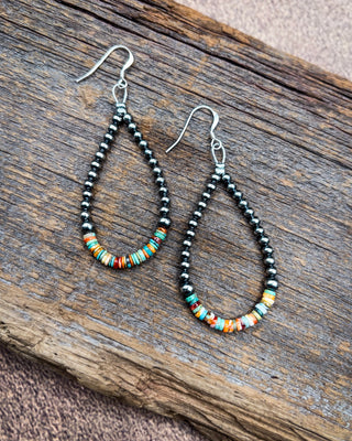 A pair of handmade teardrop dangle earrings with black seed beads and colorful striped accent beads, displayed on a rustic wooden surface.