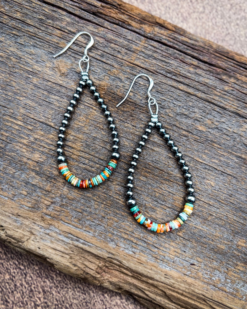 A pair of handmade teardrop dangle earrings with black seed beads and colorful striped accent beads, displayed on a rustic wooden surface.