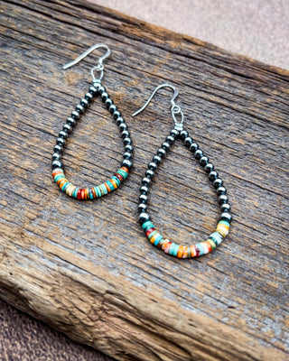 A pair of teardrop-shaped dangle earrings with black seed beads and a colorful beaded arc near the bottom, displayed on a rustic wooden surface.