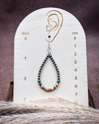 A beaded teardrop earring displayed on a wooden ear-measuring stand. The earring has dark gray beads with a small section of turquoise, orange, and white patterned beads at the bottom.