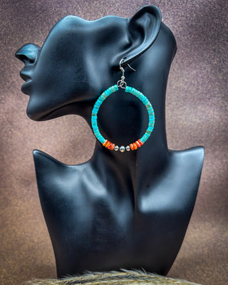 A pair of handmade hoop earrings with turquoise blue beads, orange-red accents, and small silver beads, displayed on a black female mannequin head against a textured brown background.