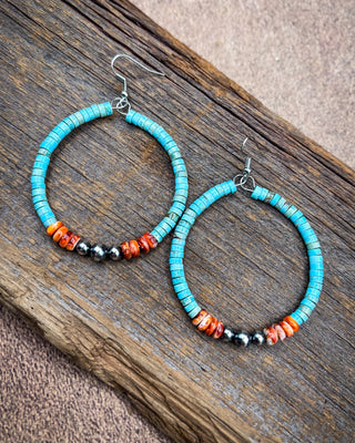 A pair of handmade hoop earrings with turquoise blue beads, orange and red marbled beads, and small black beads, displayed on a rustic wooden surface.