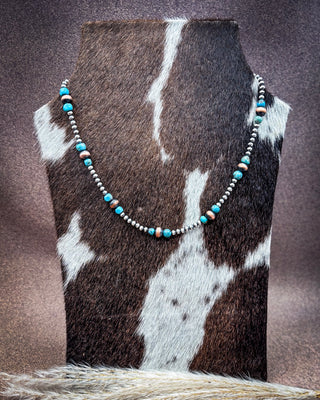 A beaded necklace displayed on a cowhide jewelry bust against a textured brown backdrop. The necklace features turquoise blue beads, small silver-toned round beads, and a few peach-orange accent beads.