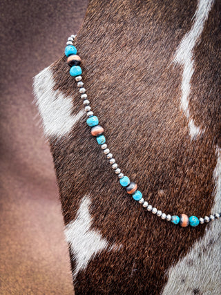 A beaded necklace draped over a brown and white cowhide surface, featuring turquoise-blue beads, small white beads, and a few peach-colored beads.