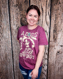 A girl modeling the Adios Cowboy Distressed Graphic Tee with a wood back drop.