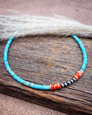 A beaded necklace with turquoise-blue beads, red-orange striped beads, and metallic silver beads, displayed on a rustic wooden surface with dried grass in the background.