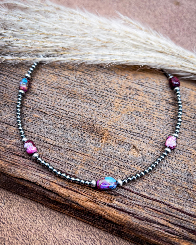 A beaded necklace with dark metallic beads, pink stones, and a central iridescent oval bead, displayed on a rustic wooden surface with dried grass.
