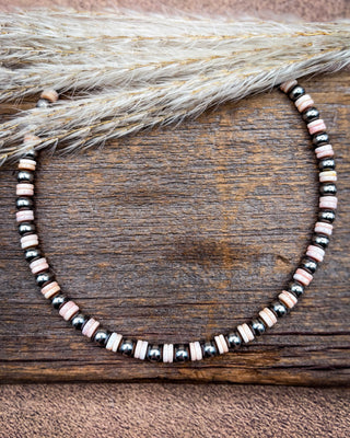A beaded necklace with alternating black metallic beads, pale pink striped beads, and natural wood slice beads, displayed on a rustic wooden surface with dried grass.