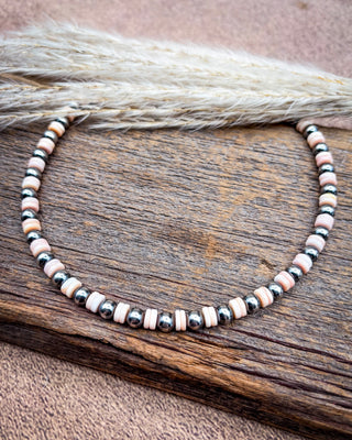 A beaded necklace with alternating light pink and black marbled beads, displayed on a rustic wooden surface with dried grass in the background.