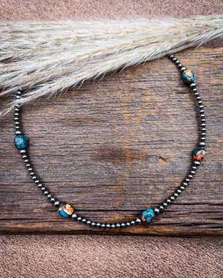A beaded necklace with dark metallic beads and turquoise, orange, and white marbled accent beads, displayed on a rustic wooden surface with dried pampas grass.