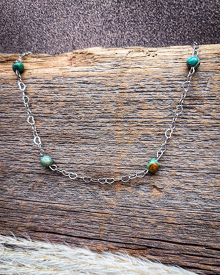 A delicate silver chain necklace with four small round turquoise beads and two brown beads, displayed on a rustic wooden surface.