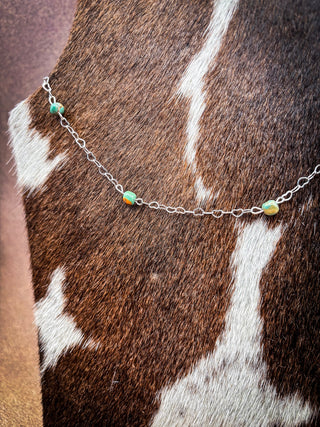 A delicate silver chain necklace with small green and orange beads, displayed on a brown and white cowhide surface.
