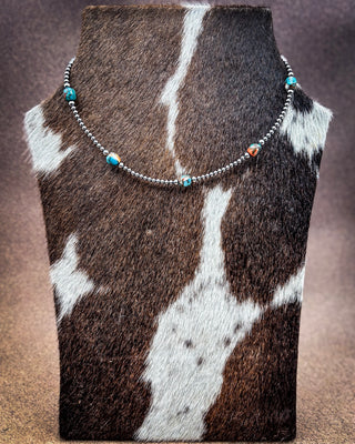 A beaded necklace displayed on a cowhide jewelry bust. The necklace features silver-toned round beads with small turquoise and orange marbled accent beads.
