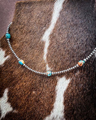 A silver beaded necklace with small turquoise and orange beads, draped over a brown and white cowhide surface.