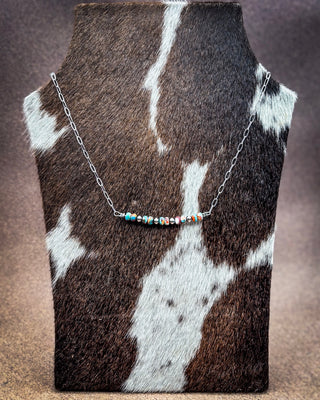 A silver chain necklace with a central cluster of small colorful beads, displayed on a cowhide jewelry bust against a brown textured background.