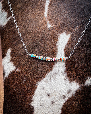 A silver chain necklace with a curved bar of colorful patterned beads, displayed on a brown and white cowhide background.