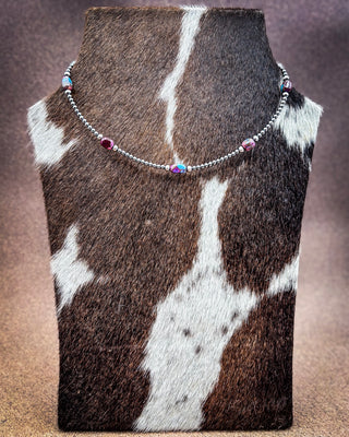 A silver beaded necklace with small iridescent beads and two dark red gemstone accents, displayed on a cowhide mannequin bust against a brown gradient background.