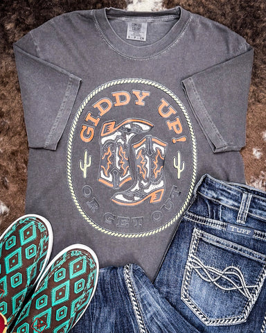 The Giddy Up or Get Out Graphic Tee displayed on cowhide along with shoes and jeans.