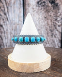 The Rayna Genuine Turquoise Bracelet on a display stand on a wood table with a wood backdrop.