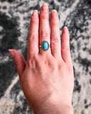 The Riley Adjustable Genuine Turquoise Ring displayed on a finger with carpet in the background.