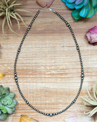 A beaded necklace made of small dark gray beads with a silver clasp, arranged in an oval shape on a light wood surface surrounded by decorative dried flowers and succulents.