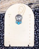 The Bitty Blue Stud Fashion Earrings displayed on display case in front of black pebbles.