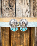 The Bitty Blue Stud Fashion Earrings displayed on wood table in front of wood backdrop.