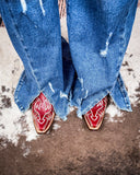 Close-up of blue jeans with a distressed look and red cowboy boots with white designs on a textured surface.