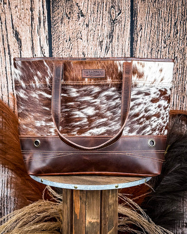 The Westin Hair on Hide Tote Bag on a wooden table with wheat in the background.
