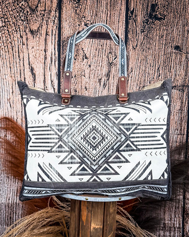 The Black Sands Weekender Bag displayed on a wooden table with wheat in the back, wood backdrop.