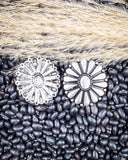 Two silver floral earrings on a bed of black beans and straw.