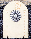 Silver concho earring on a wooden scale with measurement markings, surrounded by black beans.