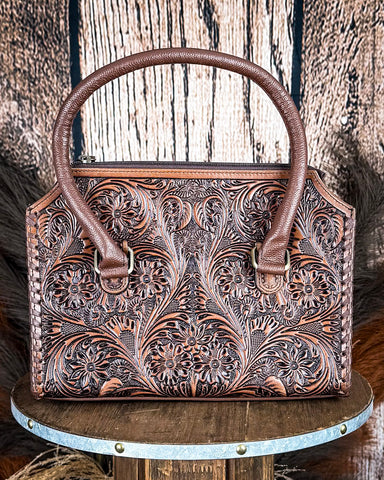 Brown leather handbag with intricate floral patterns on a wooden surface.