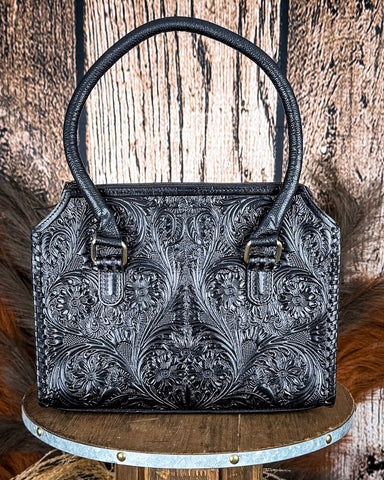 Black leather handbag with intricate patterns on a wooden stand against a rustic wooden background.
