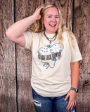 A girl wearing the Tough Luck Cowboy Graphic Tee with one hand on her leg, and the other in her hair. She is standing in front of a wooden backdrop.