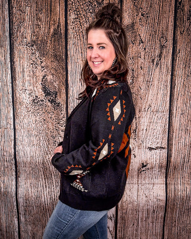 A woman standing side ways against a wood back drop, wearing the Stand by Me Knit Sweater and medium washed jeans. 