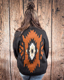 A woman facing the wood paneling behind her to show the Aztec design on the back of the sweater.