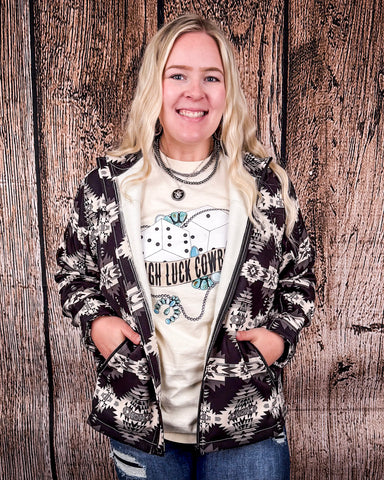 A girl wearing the Aztec City Soft Shell Jacket with both hands in her pockets. The jacket is open, showing the graphic tee underneath. She is standing in front of a wood back drop. 