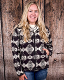 A girl is wearing the Aztec City Soft Shell Jacket, it is zipped up and she has one hand in her pocket with the other ion her leg. She is standing in front of a wood back drop.
