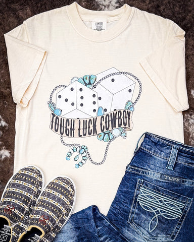 The Tough Luck Cowboy Graphic Tee displayed on cowhide along with jeans and shoes.