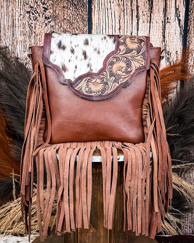 The Loralei Hair on Hide Fringe Crossbody Bag displayed on a wooden table with wheat behind it, sitting in front of a wood back drop.