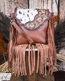 The Loralei Hair on Hide Fringe Crossbody Bag displayed on a wooden table with wheat behind it, sitting in front of a wood back drop.
