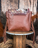 The Loralei Hair on Hide Fringe Crossbody Bag, turned around to show the back of the purse, made out of leather with a zipper on the top. It is being displayed on a wooden table with wheat and a wood back drop behind it.